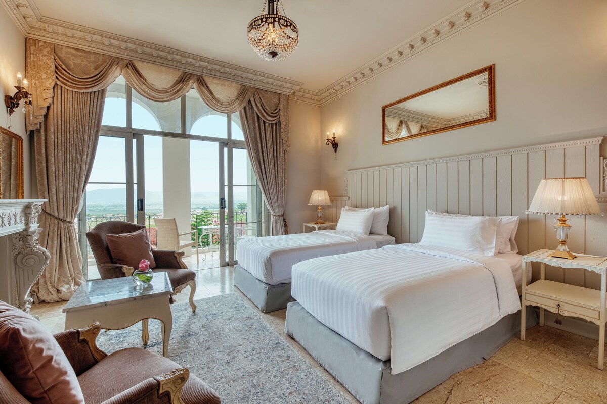 Une chambre d’hôtel avec deux lits, un coin salon comprenant une chaise et une table, un lustre, une grande fenêtre avec des rideaux et une vue sur l’extérieur.