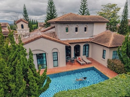 Una casa grande con piscina, rodeada de árboles y otras viviendas.