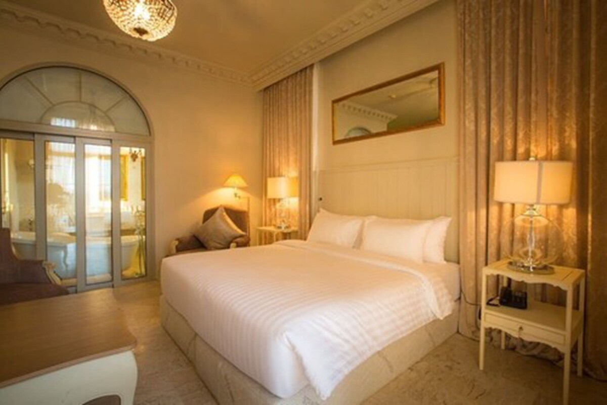 Une chambre d’hôtel avec un grand lit, deux tables de chevet avec des lampes, un miroir au-dessus du lit et une vue sur une piscine, visible à travers une porte vitrée.