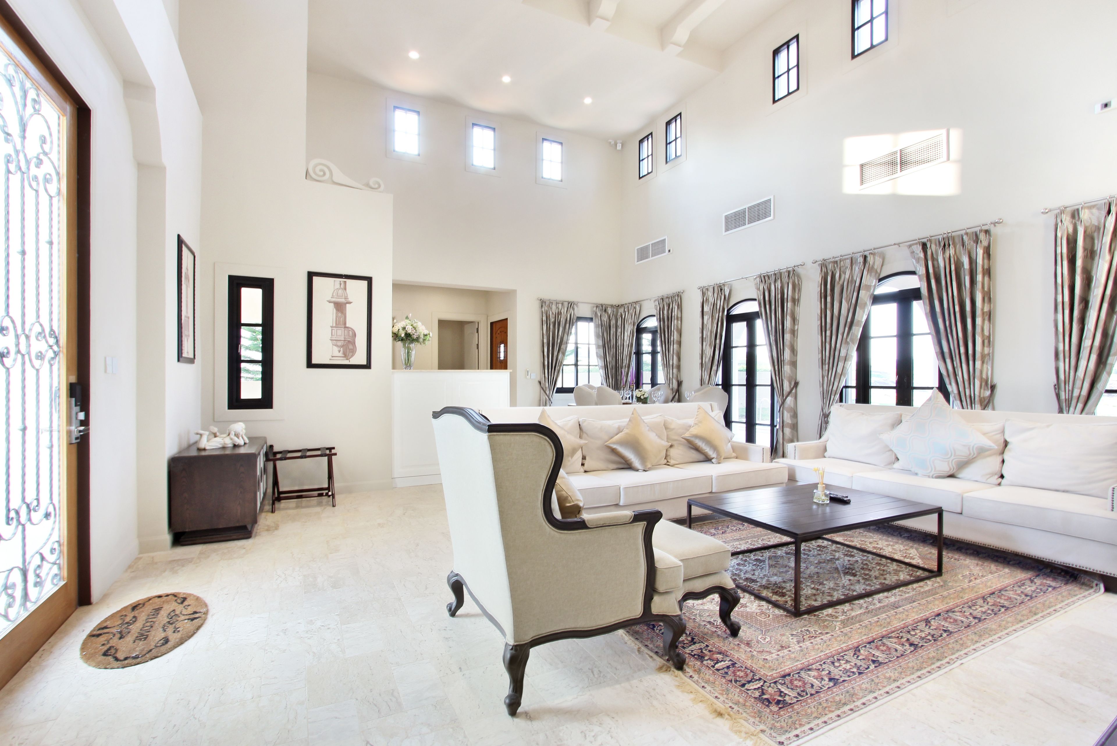 A spacious living room with a large sofa, a coffee table, and a decorative chair. There are multiple windows with curtains, a fireplace, and framed artwork on the walls.