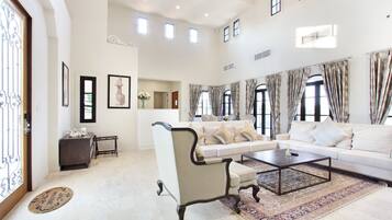 A spacious living room with a large sofa, a coffee table, and a decorative chair. There are multiple windows with curtains, a fireplace, and framed artwork on the walls.