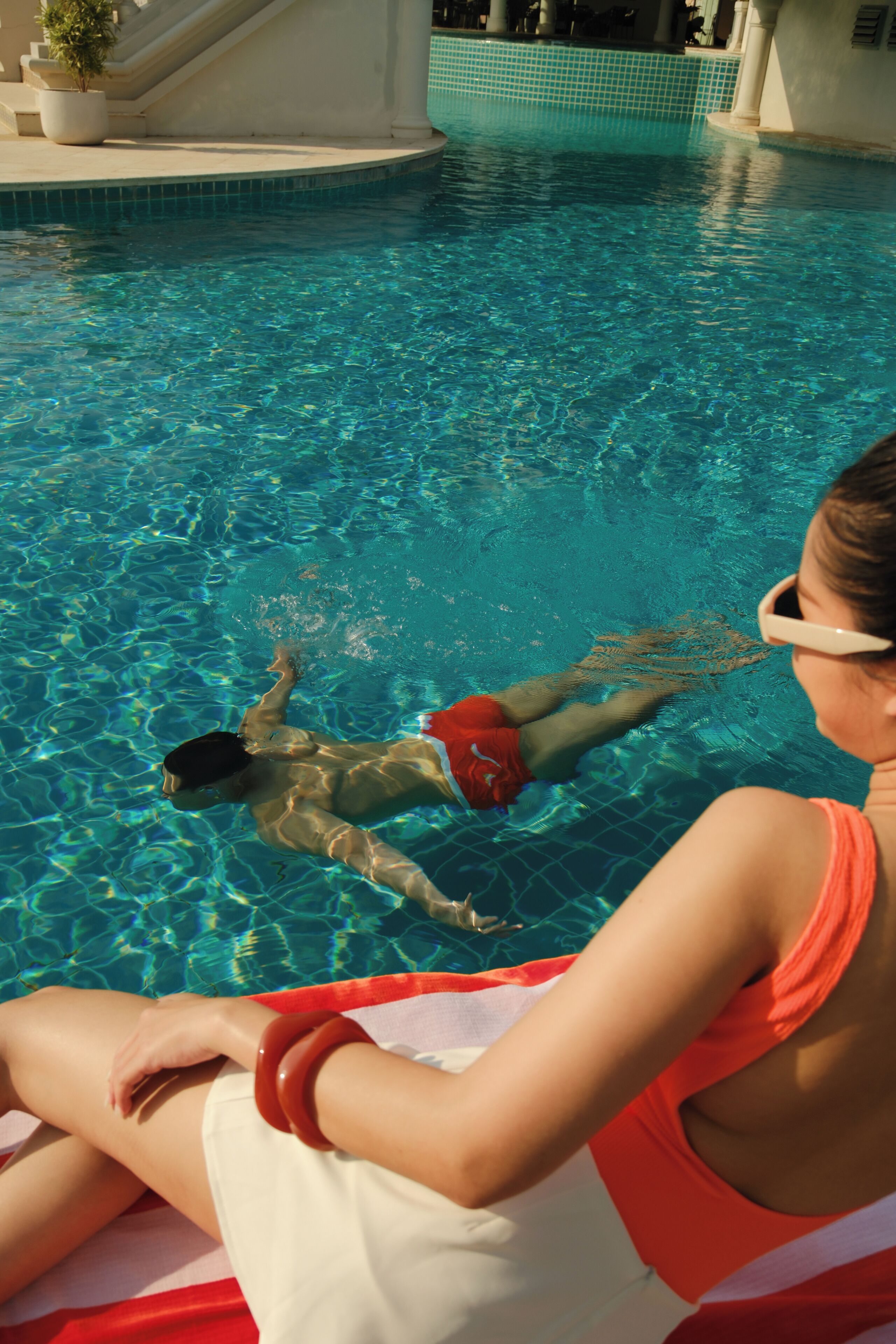 A swimming pool with clear water, a person sitting on a lounge chair, and a dog in the water.
