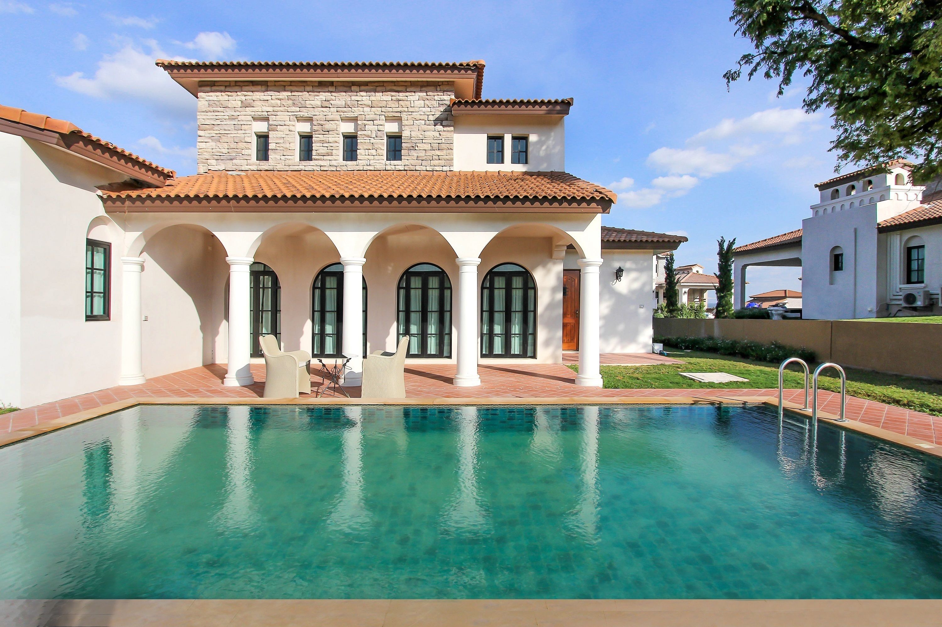 Vista a la piscina de una casa de dos pisos, con piscina, columnas blancas y techo de tejas.