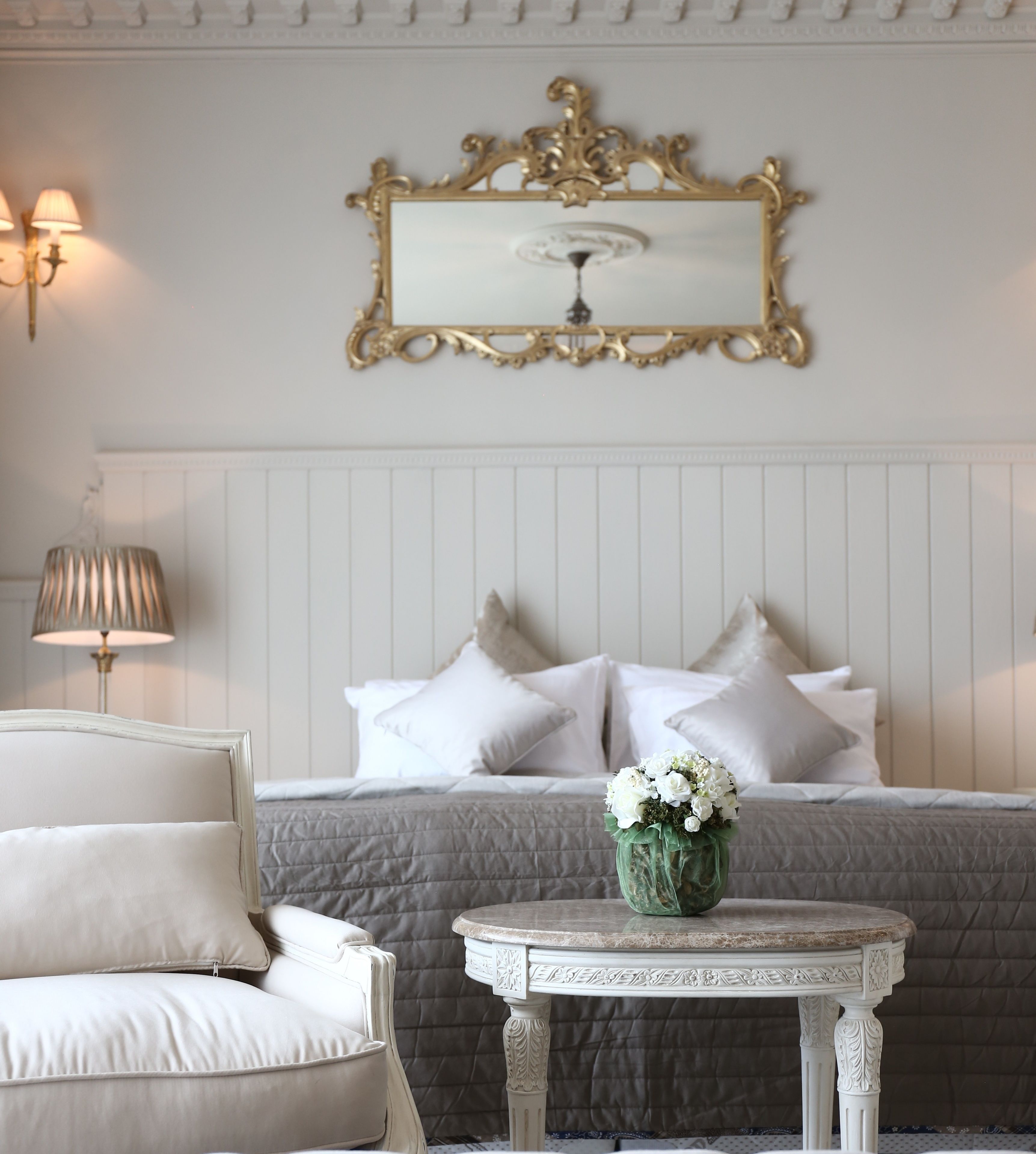 A neatly arranged bedroom with a bed, pillows, a bedside table with a vase, and a wall-mounted mirror.