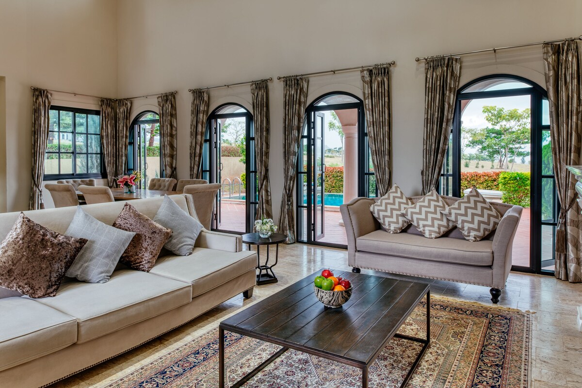 Un salon spacieux avec un canapé beige, une table basse sur laquelle se trouve un panier de fruits, et une vue sur un jardin extérieur, visible à travers de grandes fenêtres en arc de cercle.