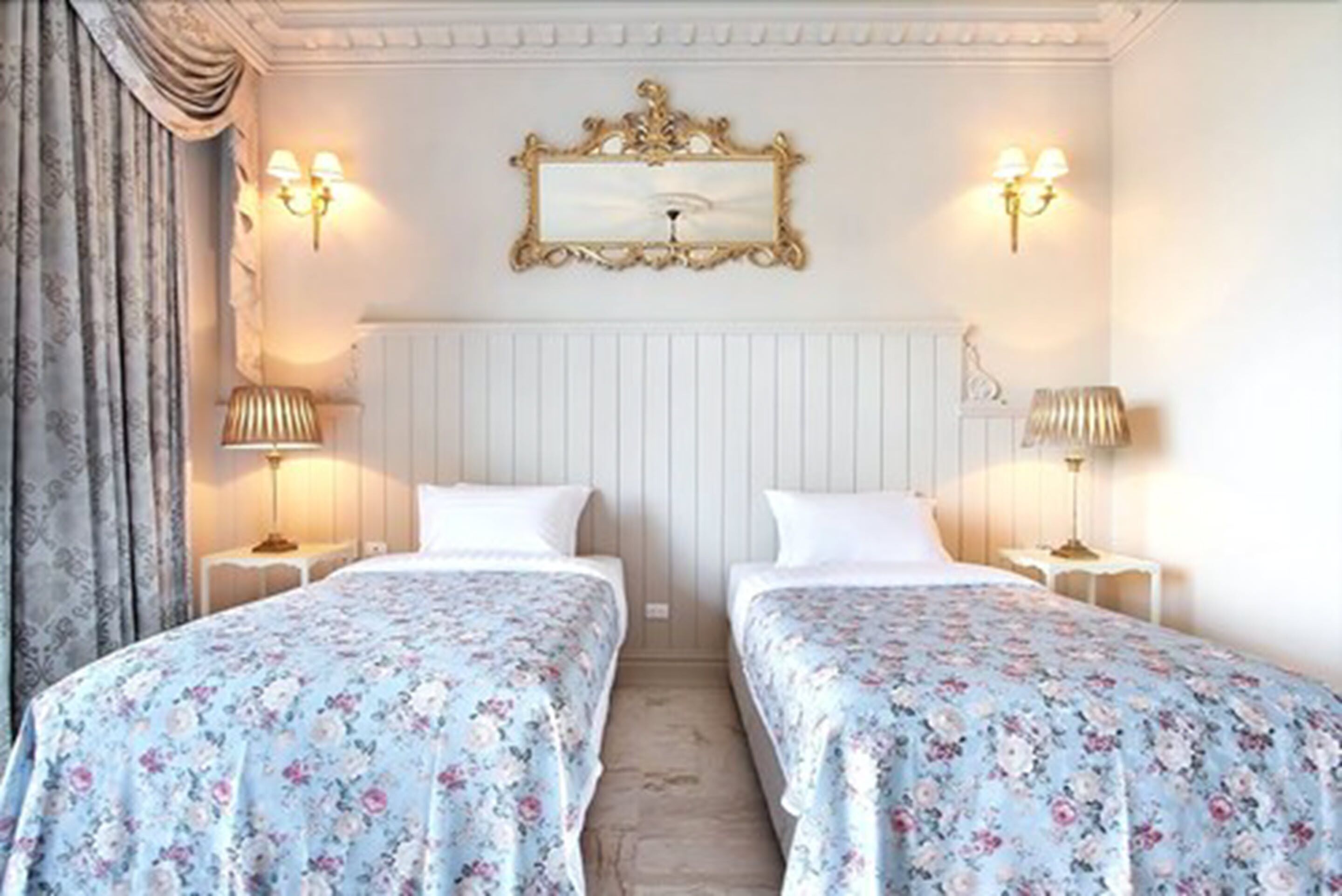 A hotel room with two beds, floral bedding, a white radiator, and wall-mounted lamps.