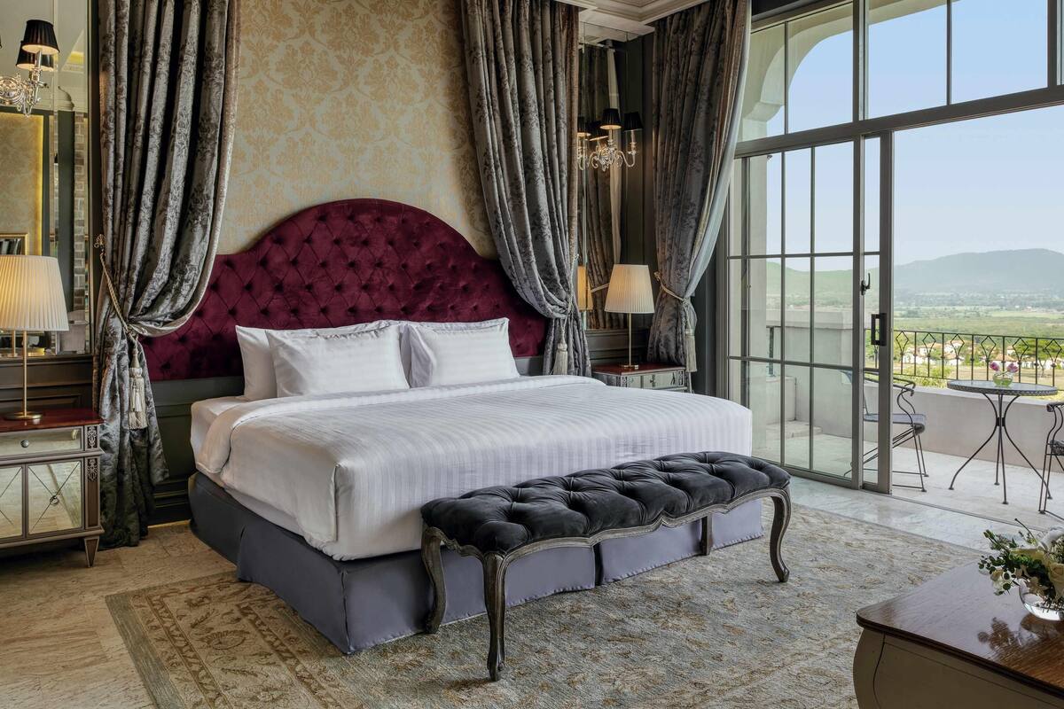 A bedroom with a large bed, a bench, a nightstand, a lamp, and a view of the outdoors.