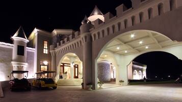 A grand castle-like building with a large entrance, a tower, and a driveway with vehicles.