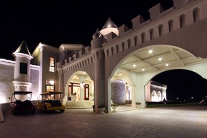 A grand castle-like building with a large entrance, a tower, and a driveway with vehicles.