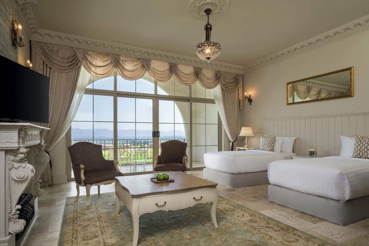 A hotel room with a large window, two beds, a TV, and a chandelier.