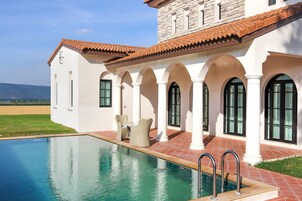 A poolside area with a clear view of the pool, a patio with outdoor furniture, and a stone building with arches.