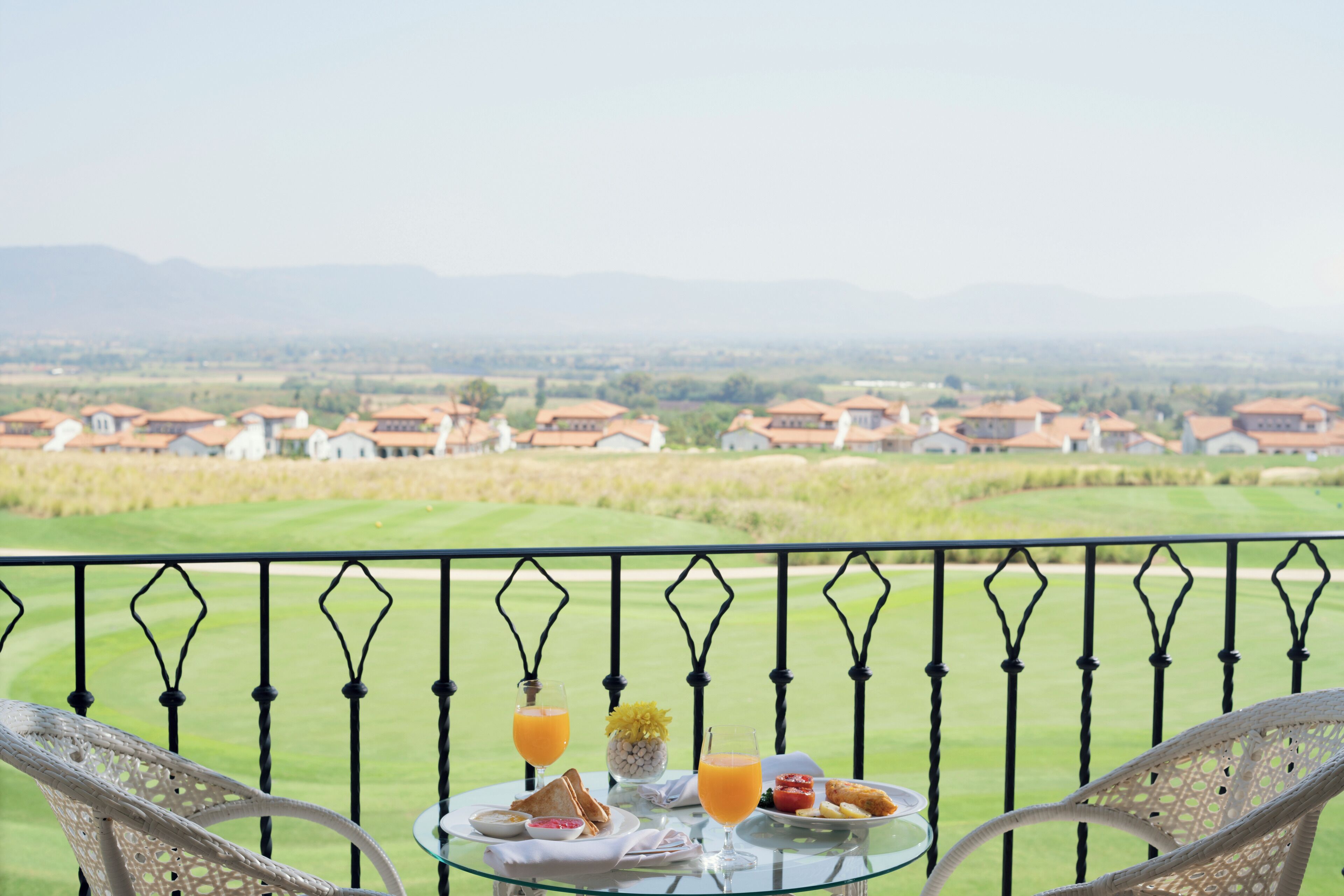 Una mesa con alimentos para el desayuno y un vaso de jugo de naranja en un balcón con vista a un campo de golf y edificios residenciales.