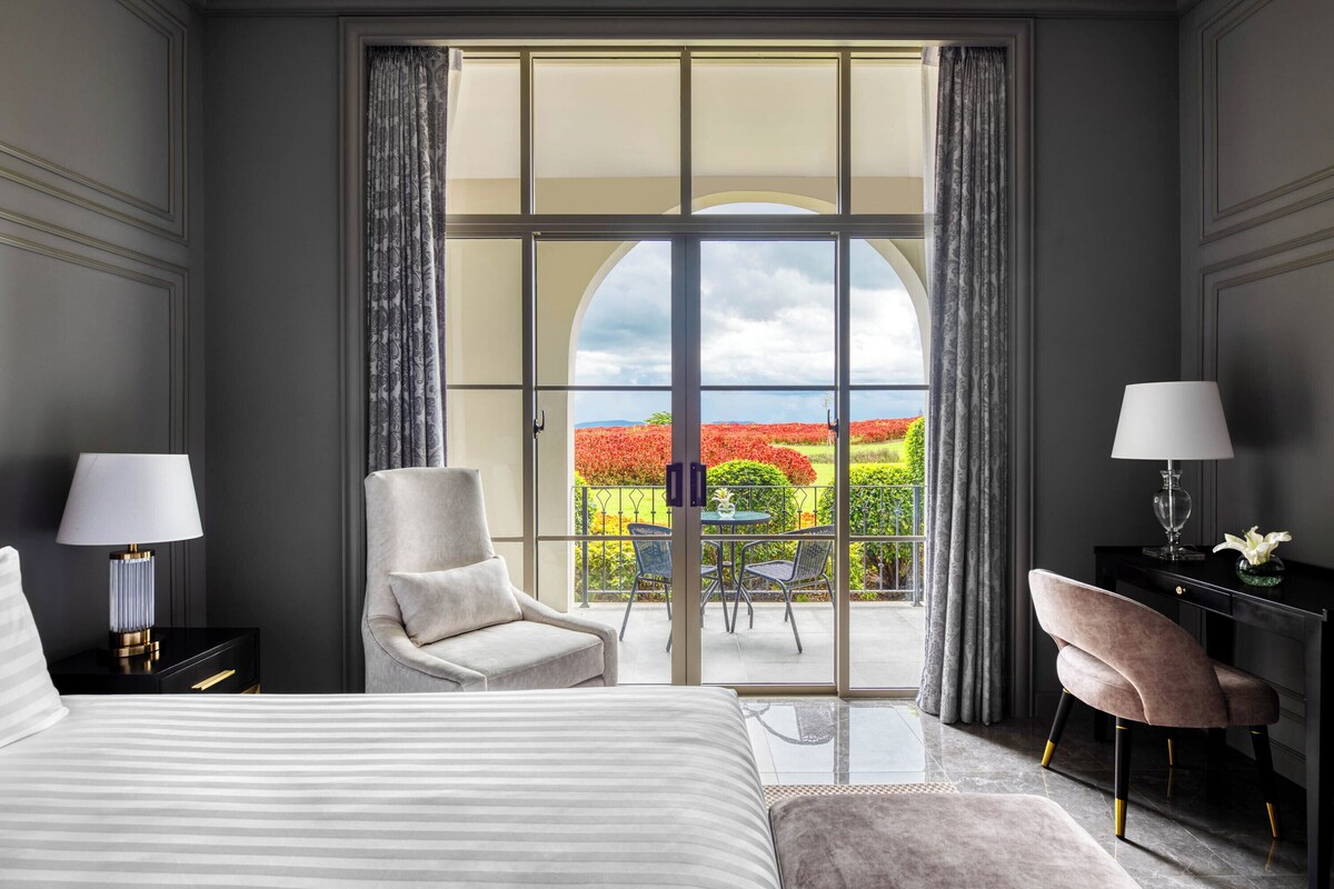 Une chambre d’hôtel avec un grand lit, deux fauteuils, un bureau et une vue sur un jardin, accessible par une porte vitrée.