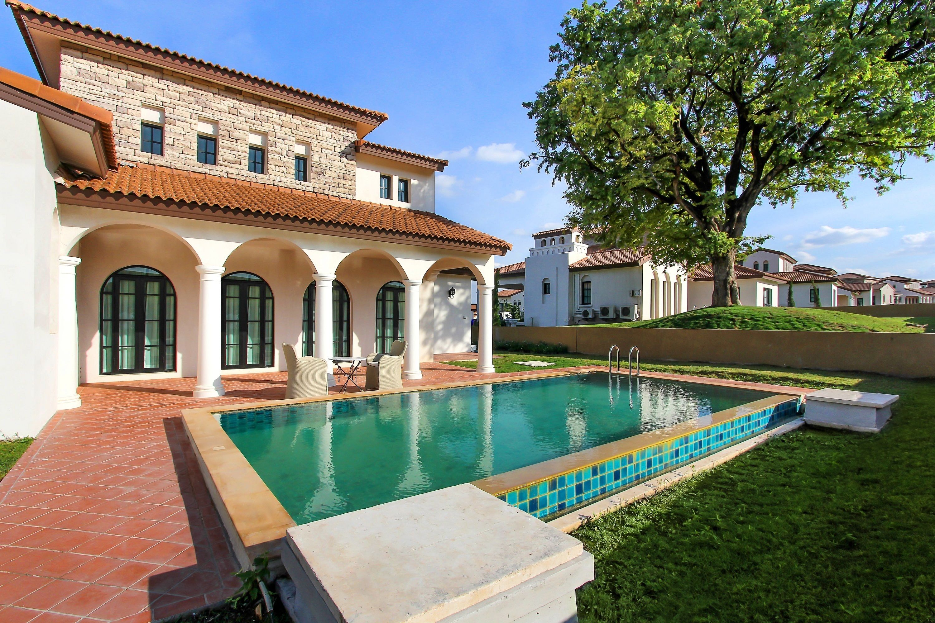 A poolside view of a house with a tiled patio, a swimming pool, and a large tree.