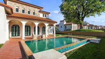 A poolside view of a house with a tiled patio, a swimming pool, and a large tree.