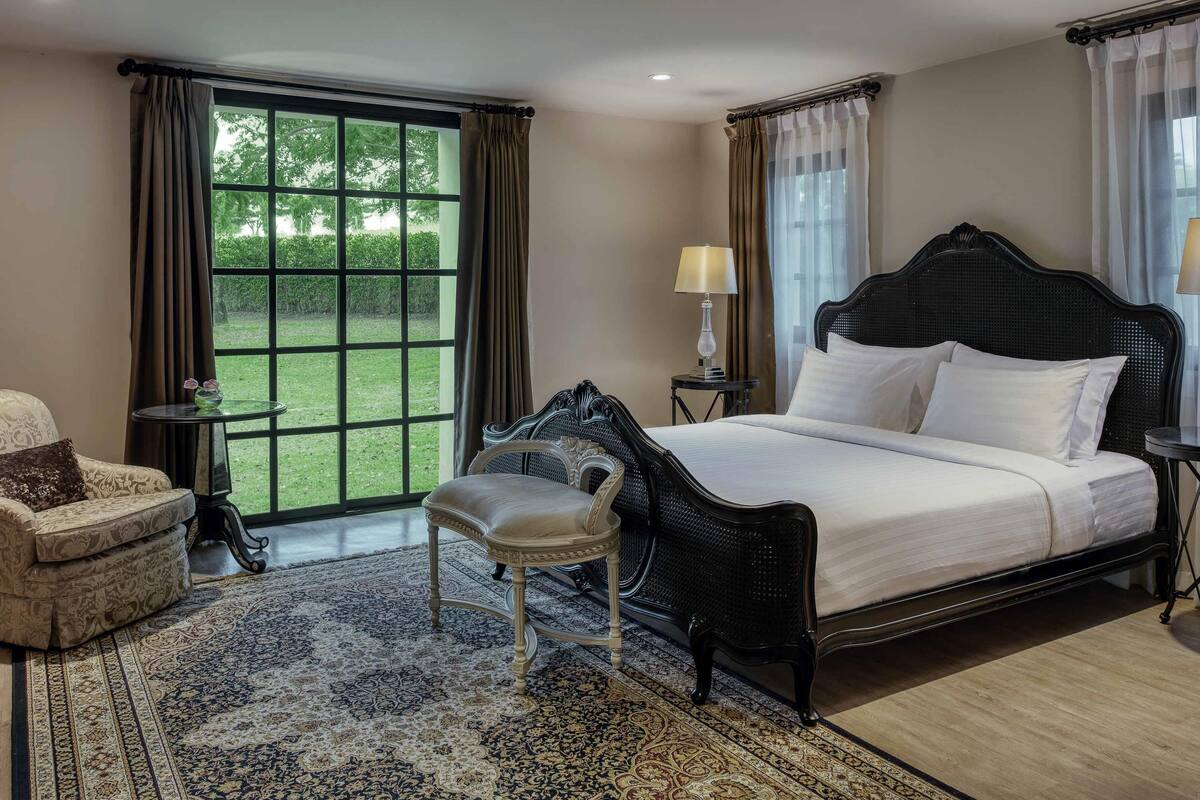 A bedroom with a large bed, a chair, a small table, and a view of a green garden through a window.