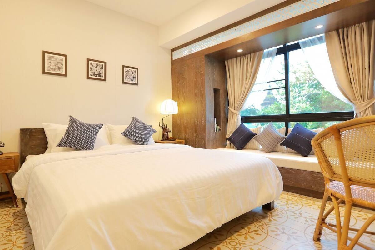 A hotel room with a large bed, a bedside table with a lamp, a chair, and a window with curtains.