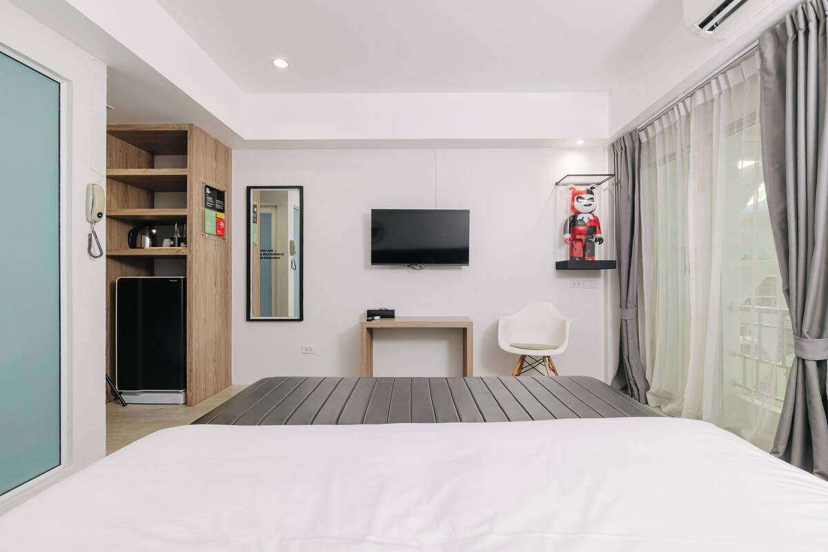 A modern hotel room with a bed, a TV, a desk, a chair, a wardrobe, and a window with curtains.