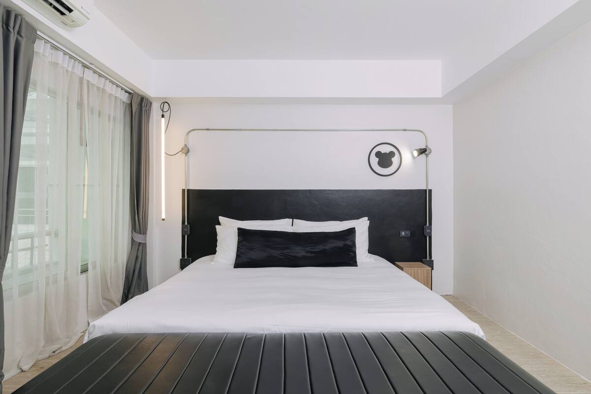A modern hotel room with a large bed, a headboard with a circular logo, a bedside table, and a window with curtains.