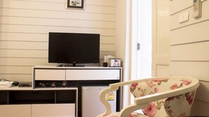 Habitación con televisión de pantalla plana sobre un mueble blanco, un refrigerador pequeño, una silla estampada y una puerta con panel de seguridad.