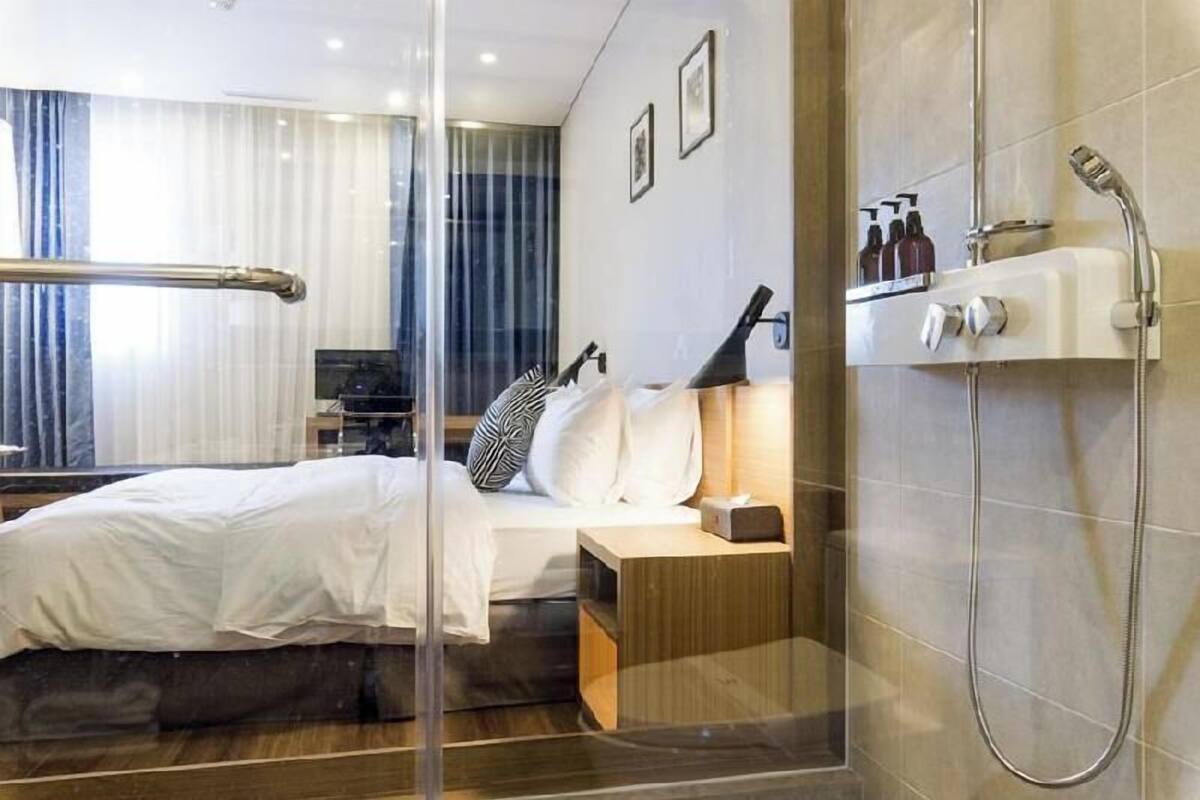 Une chambre d’hôtel moderne avec une cloison en verre, un lit, une table de chevet avec une lampe, un espace douche et une vue sur l’extérieur.