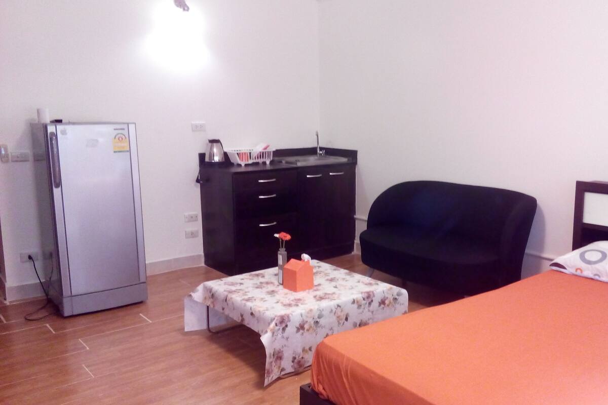 Ein kleines, schlichtes Zimmer mit einem Bett, einem Sessel, einem Schreibtisch und einem Kühlschrank.