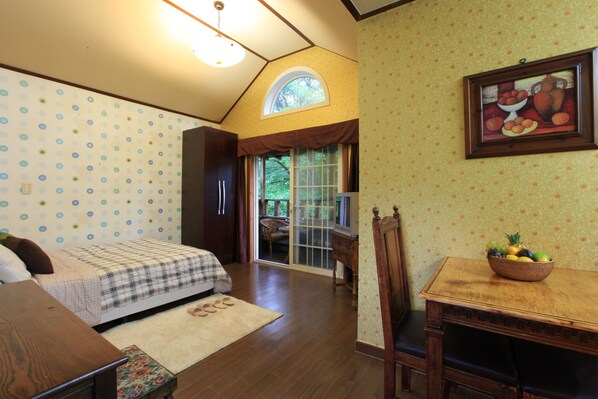 A bedroom with a bed, a wooden table with a fruit bowl, a window with a view of greenery, and a door leading to a balcony.