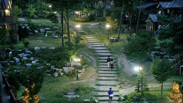 A residential area with a stone pathway leading to houses, surrounded by greenery and illuminated by street lamps. - Mayfair Pension (Pyeongchang)