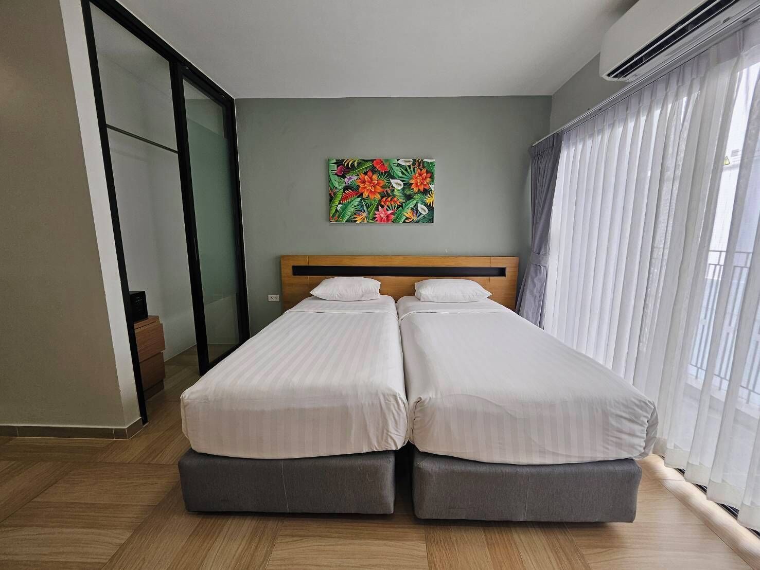 Une chambre d’hôtel avec deux lits simples, une grande fenêtre avec des rideaux légers, un tableau sur le thème des tropiques accroché au mur et une porte-fenêtre coulissante.