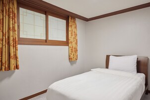 Una cama individual con cabecera de madera en una habitación con paredes blancas lisas y un ventanal con marcos de madera.