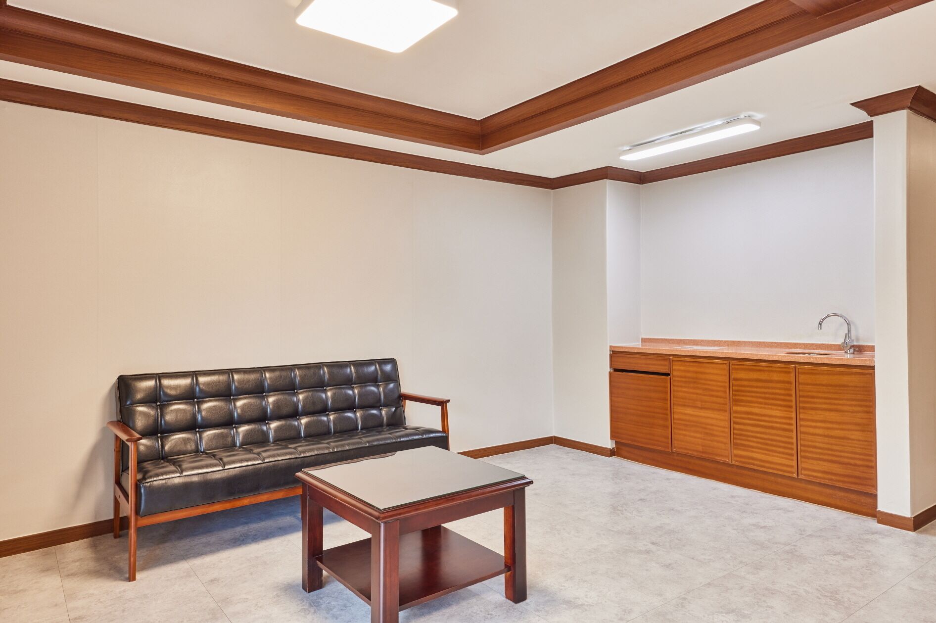 Una habitación con sofá de cuero negro, una mesa de centro de madera, un mueble de madera con lavamanos y un techo con vigas de madera e iluminación empotrada.