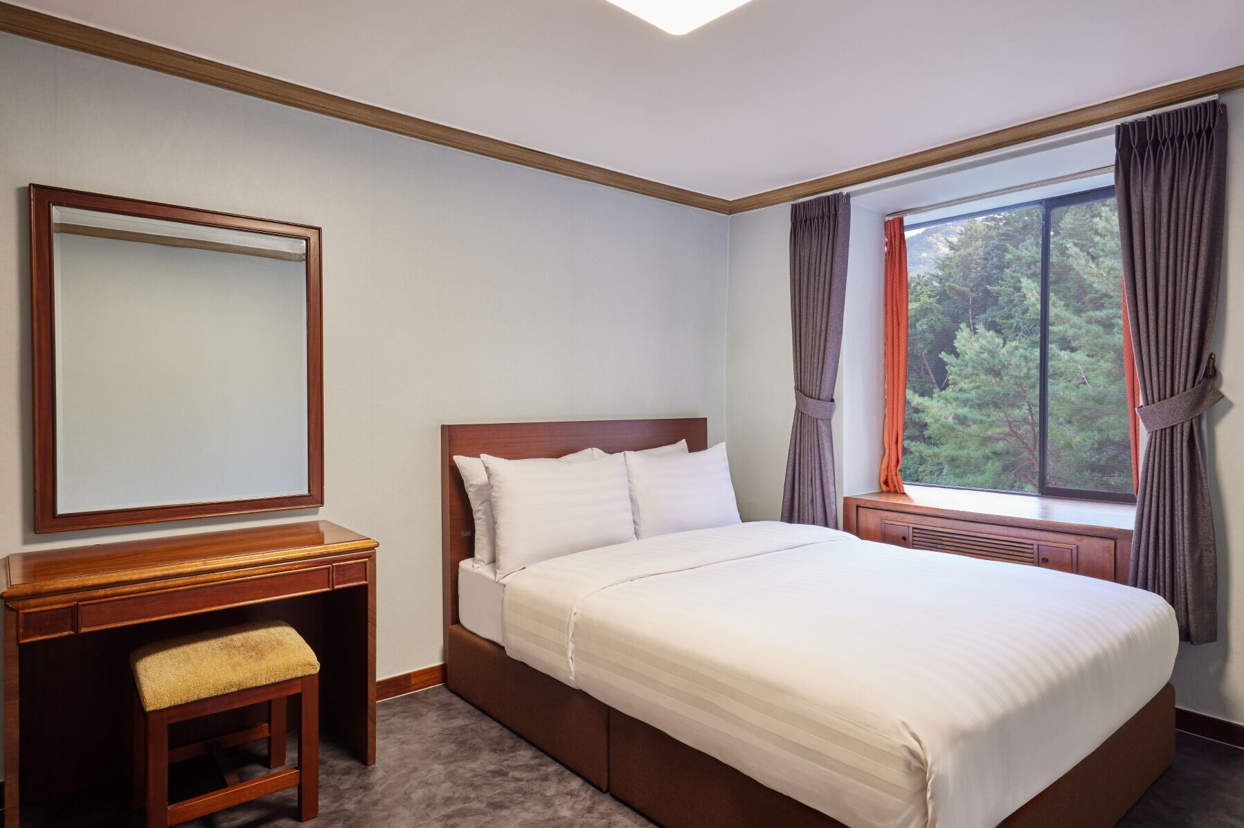 Ein Hotelzimmer mit einem großen Bett, einem Nachttisch aus Holz, einer Bank, einem Spiegel und einem Fenster mit Vorhängen.