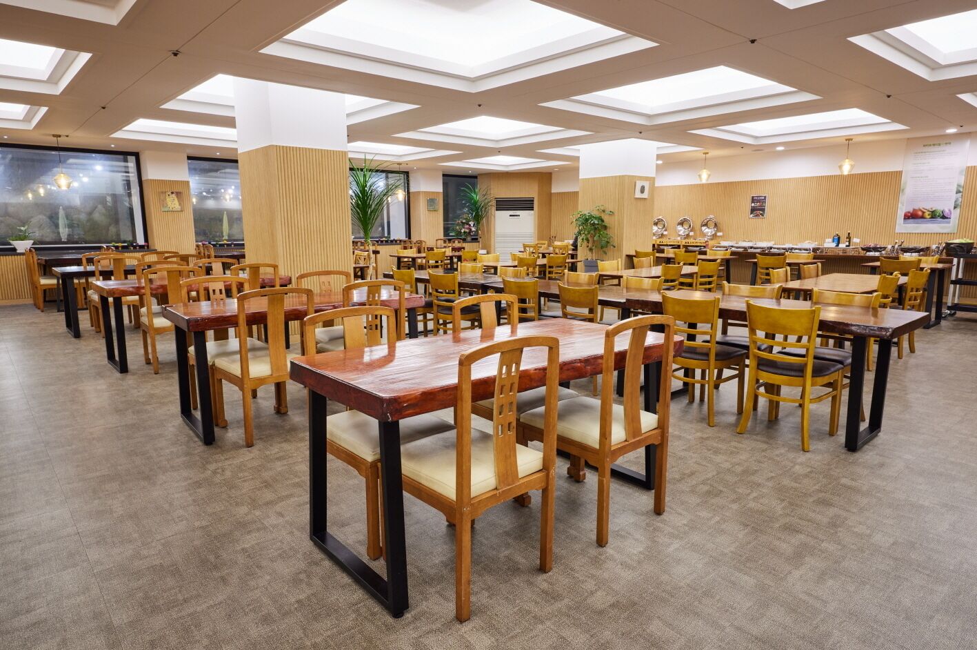 A spacious dining area with multiple wooden tables and chairs, large windows, and a high ceiling with skylights.