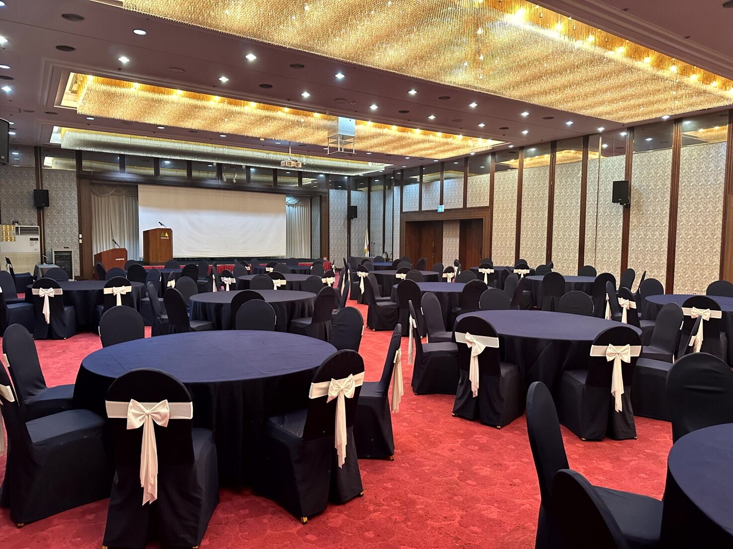A conference room with round tables, black chairs with white sashes, a red carpet, and a large screen at the front.