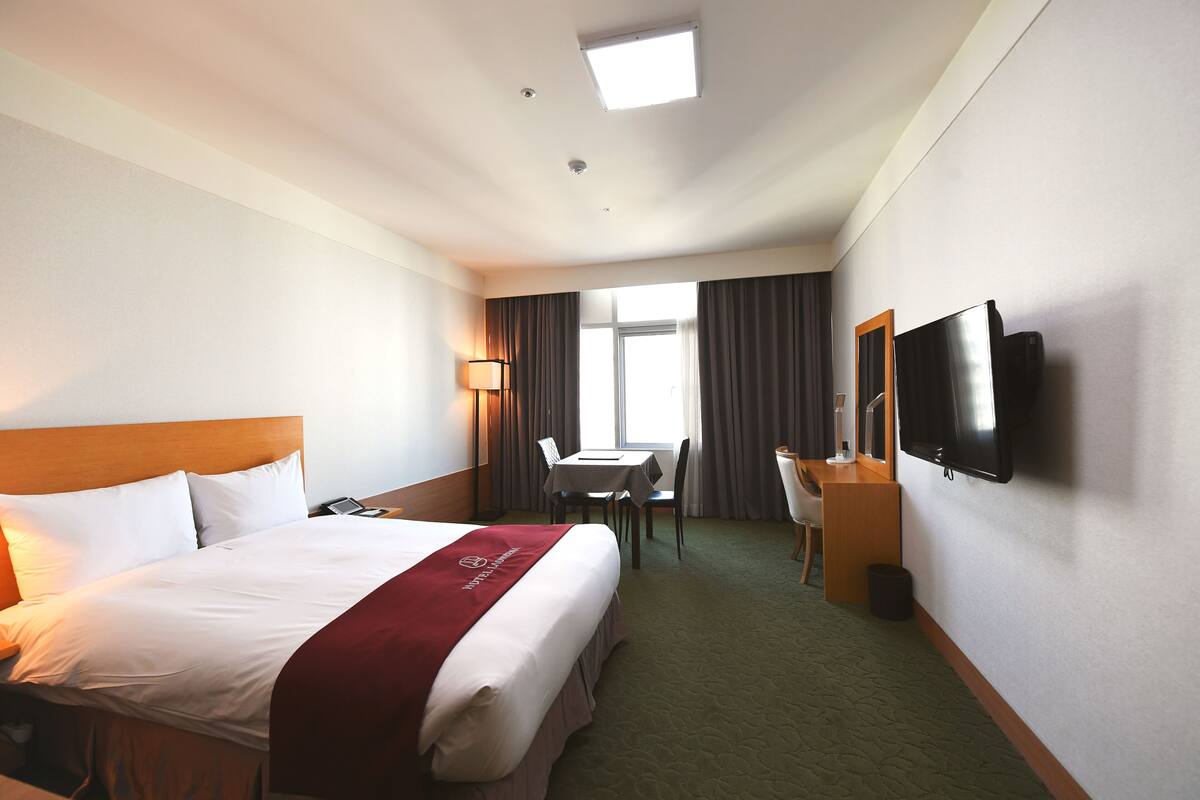 A hotel room with a large bed, a desk, a chair, a television, and a window with curtains.