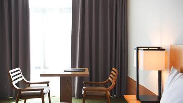 A hotel room with a bed, two wooden chairs, a small table, a lamp, and a view through curtains.