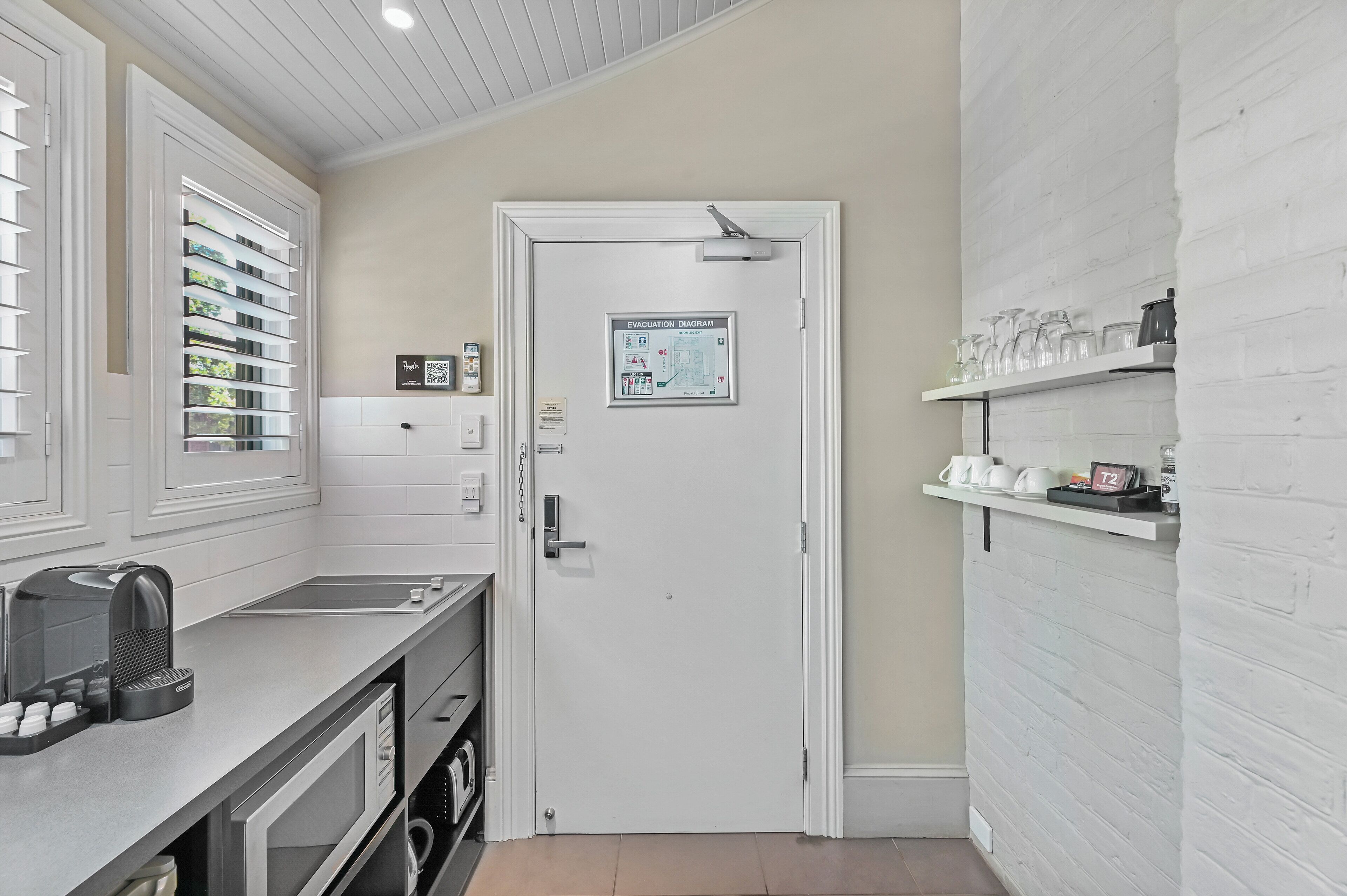 A modern kitchen with a white brick wall, a white door with an evacuation diagram, and shelves with kitchenware.