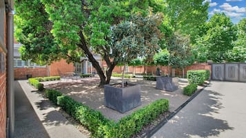 Ein Innenhof mit einem großen Baum, einer Sitzgelegenheit und einem Backsteinhaus.