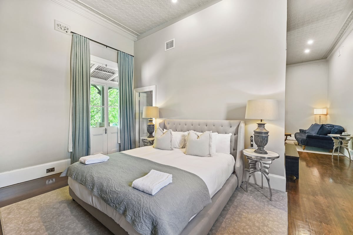 A spacious bedroom with a large bed, a bedside table with a lamp, and a view of the outdoors through a window with curtains.