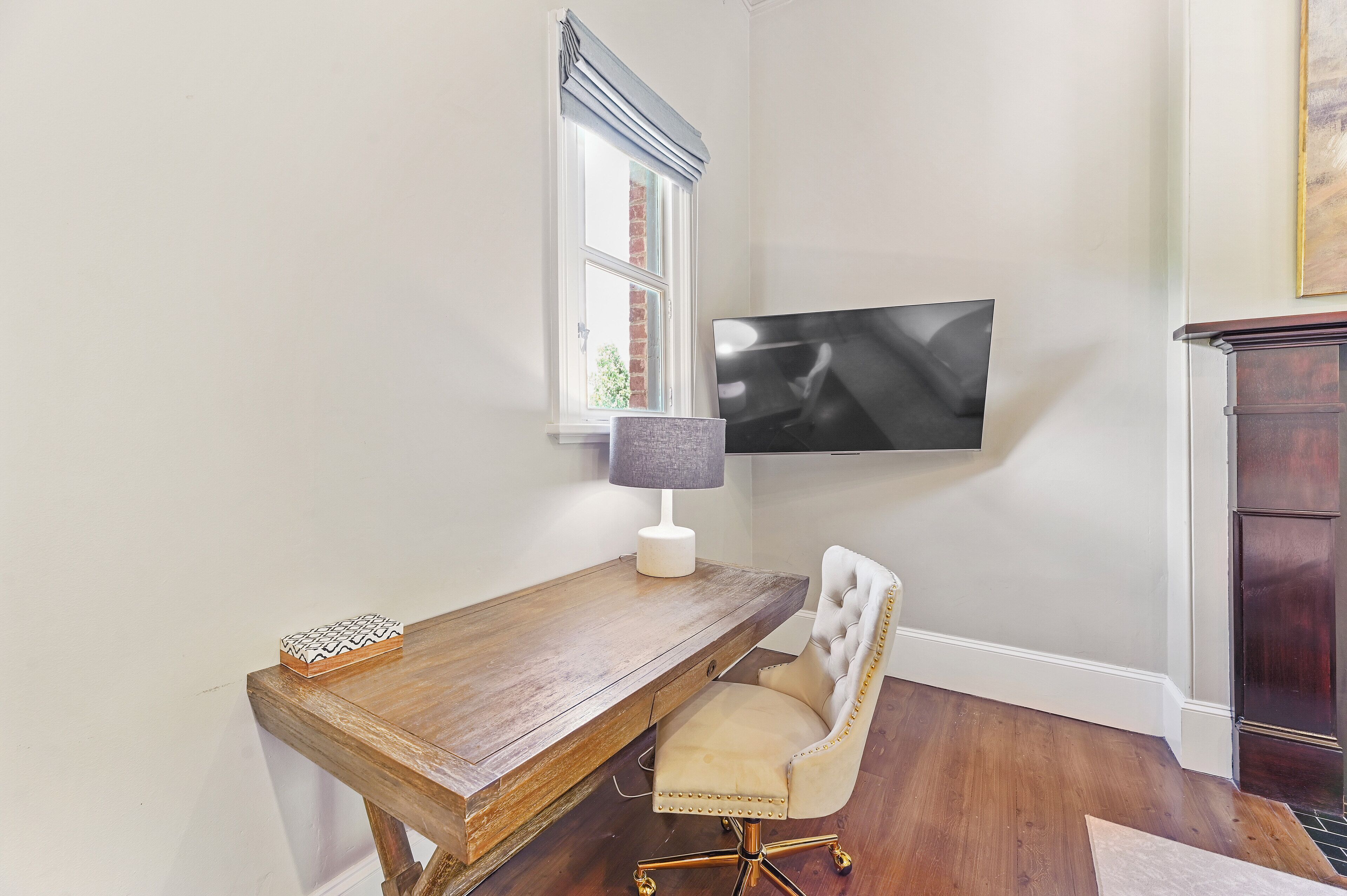 Ein Zimmer mit Schreibtisch aus Holz, einem gepolsterten Sessel, einer Lampe, einem an der Wand montierten Fernseher und einem Fenster mit Blick auf ein Backsteingebäude.