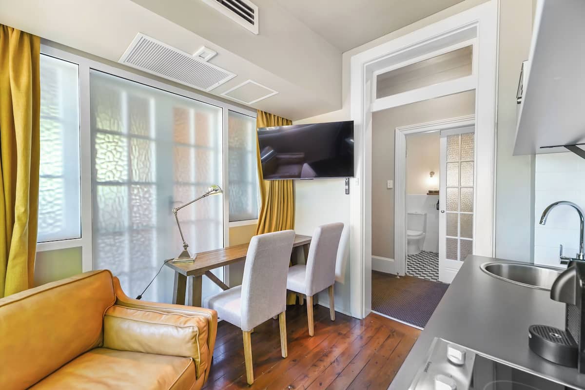 A compact living space with a sofa, dining table, chairs, a TV mounted on the wall, a kitchen area with a sink, and a bathroom visible through an open door.