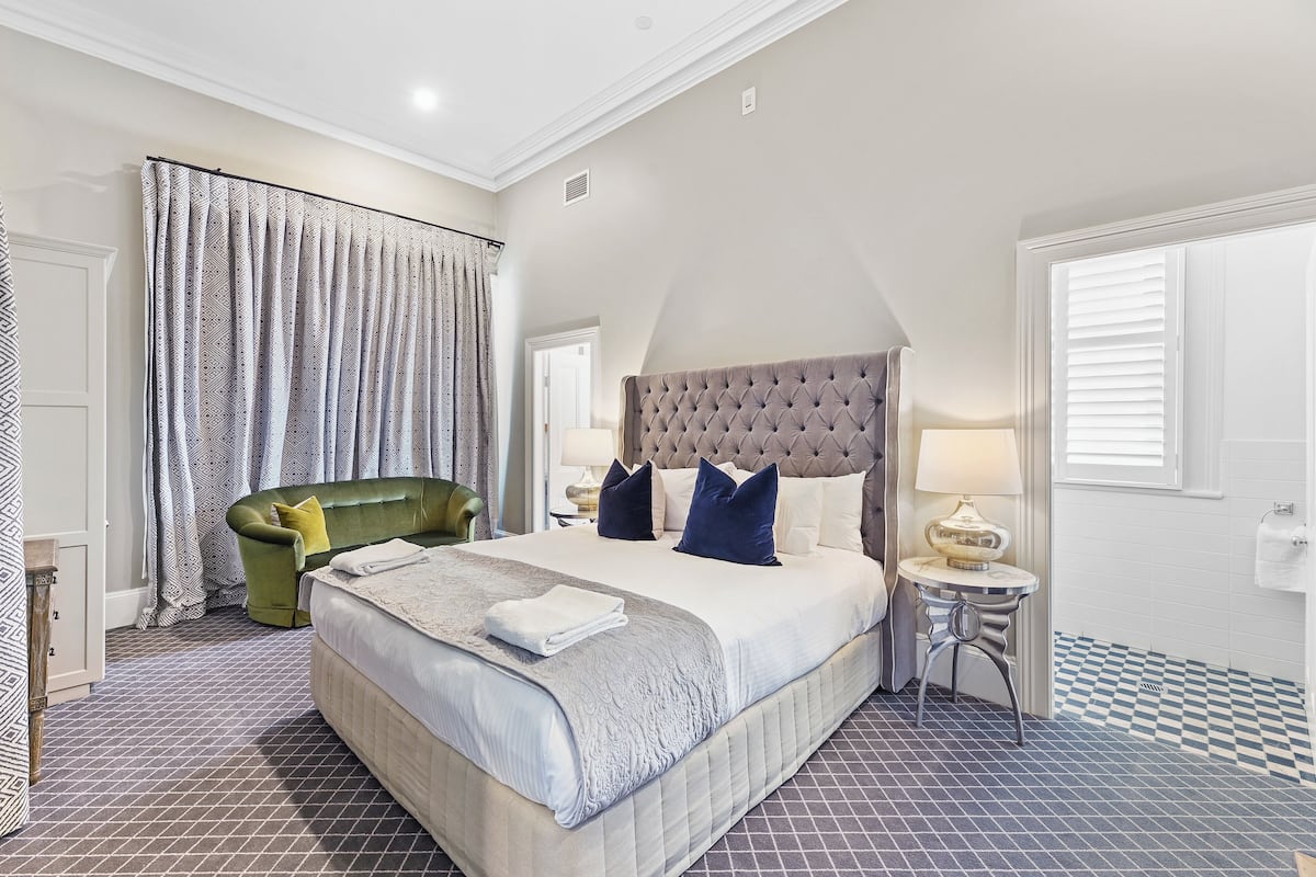 A bedroom with a large bed, a green armchair, a bedside table with a lamp, and a checkered floor.