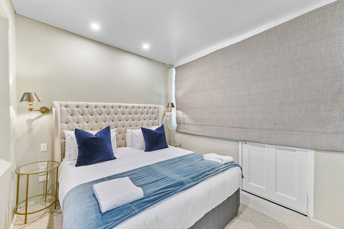 A neatly made bed with a tufted headboard, a blue and white bedspread, and a folded white towel. There are two bedside lamps with gold bases and white shades.