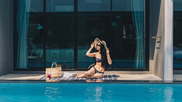 Una mujer sentada junto a la piscina, con una bebida y un bolso, frente a un edificio moderno con ventanas grandes.