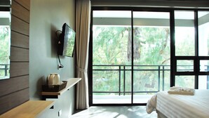 Habitación de hotel con un gran ventanal corredero a un balcón, un televisor de pantalla plana montado en la pared, una mesita de noche con hervidor y una cama bien hecha.