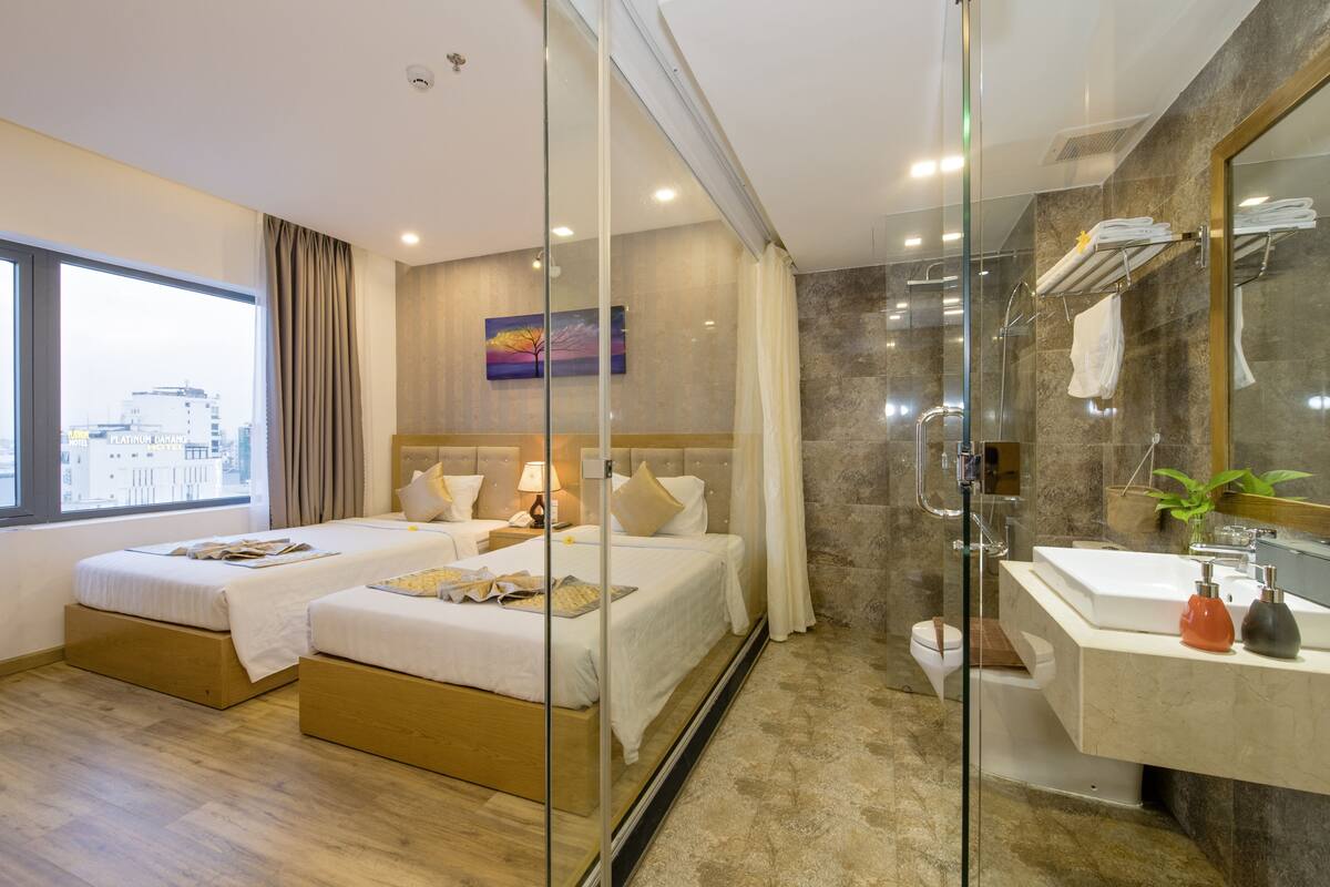 Una habitación de hotel moderna con dos camas, cabina de ducha de vidrio, baño de mármol y vistas a la ciudad.