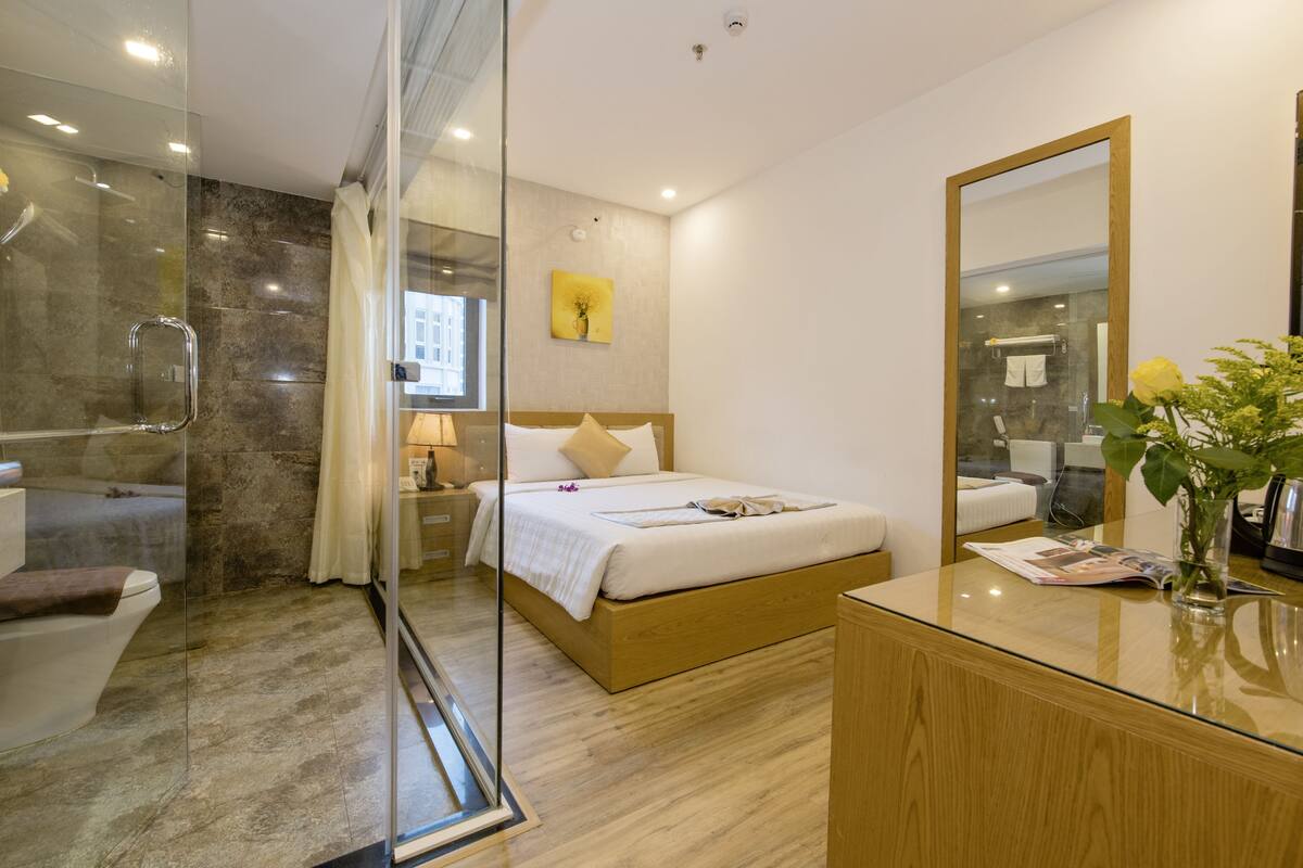 Una habitación de hotel moderna con cabina de ducha de vidrio, cama de madera, cómoda de madera y un espejo grande.