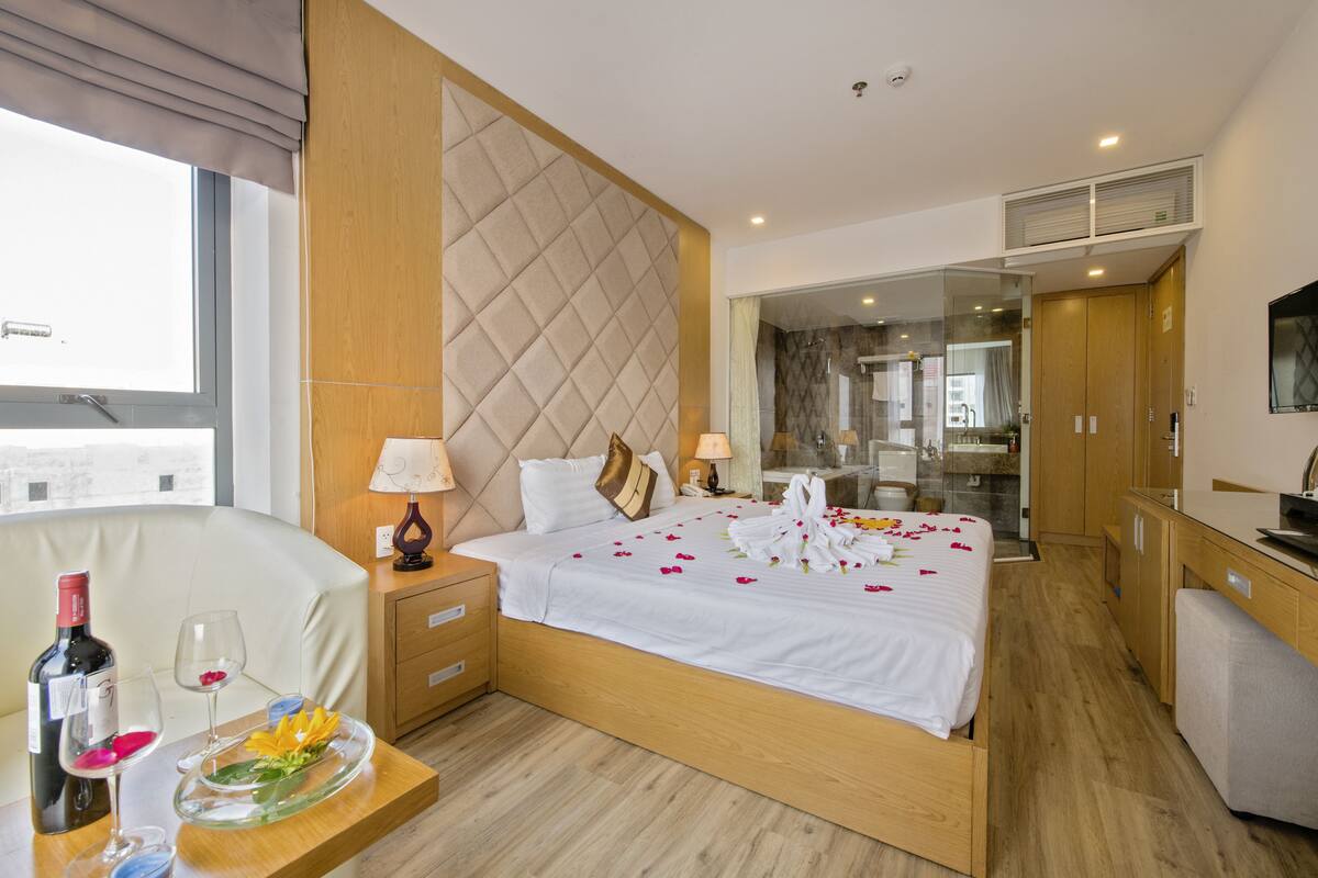 Habitación de hotel con una cama grande, mesitas de noche, un escritorio con una silla, un baño visible a través de una puerta de cristal y vistas a la ciudad.