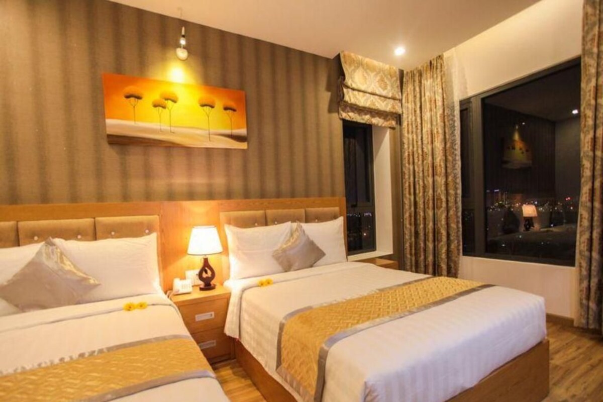 Quarto de hotel com duas camas, uma mesa de cabeceira com abajur, um quadro na parede e uma janela com cortinas.