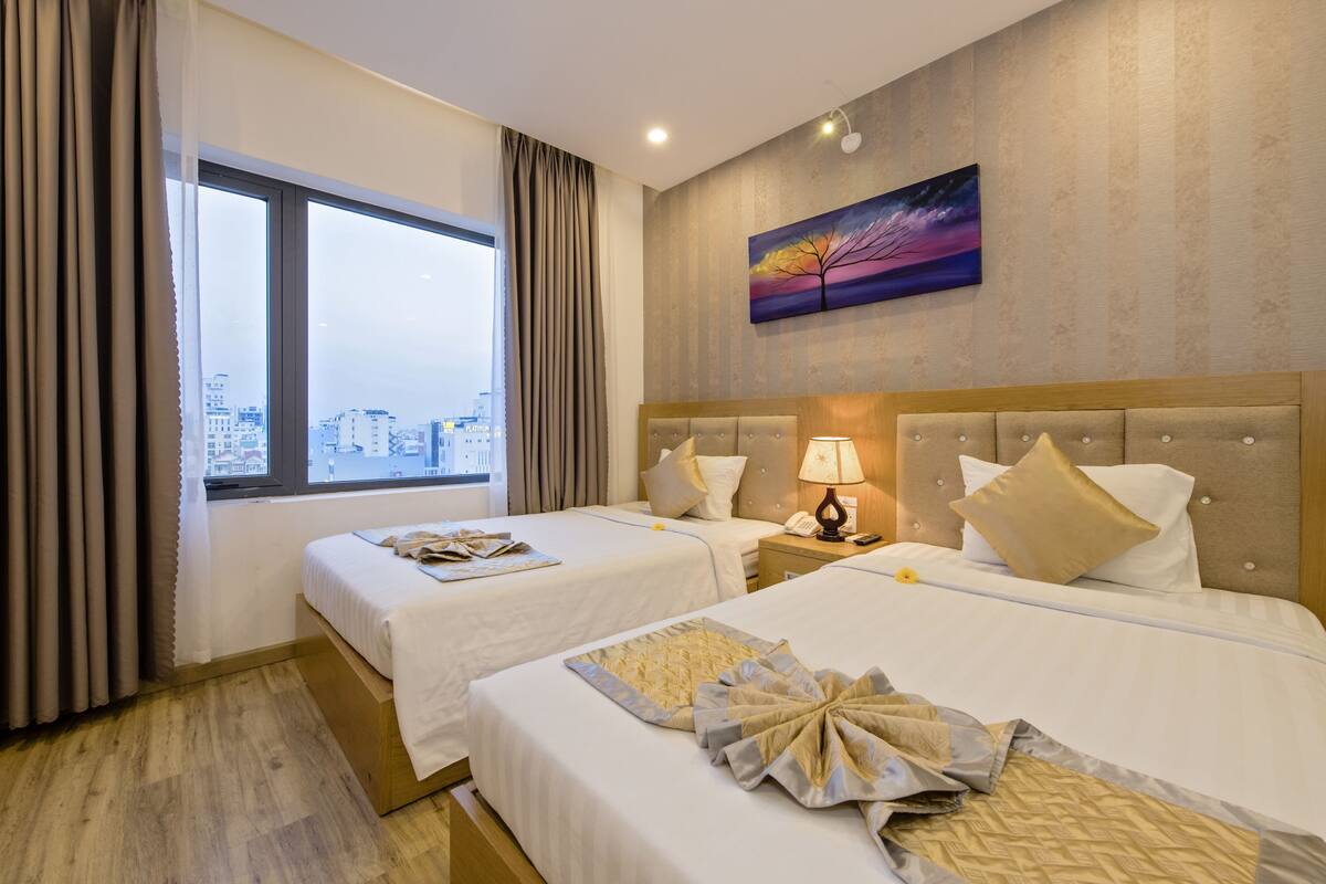 Habitación de hotel con dos camas, vistas a la ciudad, un cuadro y una mesita de noche con lámpara.
